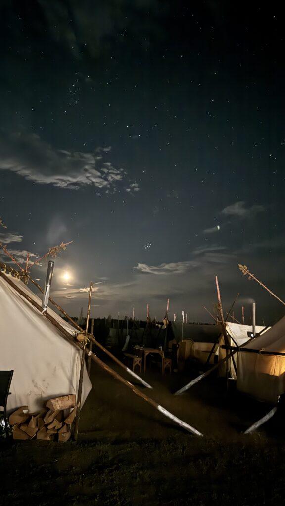 The moon shines brightly in the night sky, surrounded by stars. Tents, supported by tree branches, are dimly lit from within, offering a peaceful scene against the vast Northern sky.