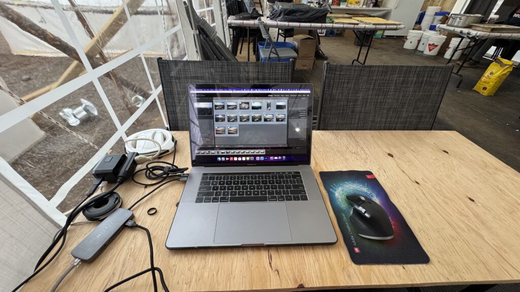 A laptop is set up on a piece of plywood balanced between two chairs in a makeshift workspace. The screen shows a photo-editing software interface, with multiple images displayed. A mouse and mousepad sit to the right of the laptop, while cables and a power adapter are visible to the left. The scene is set in the Naskapi Kitchen, with folding chairs and tables in the background. The setting feels temporary and improvised, yet functional.