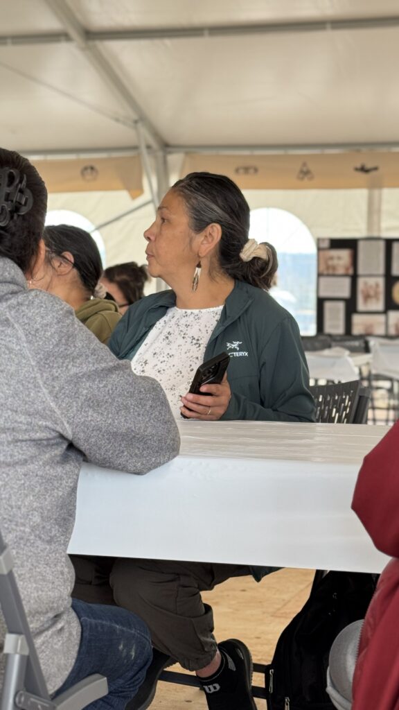 A woman with gray-streaked hair is sitting at a white table inside a large tent, wearing a green Arc'teryx jacket and holding a phone in her right hand. Her expression is thoughtful as she looks to the left. Other people are seated at the table around her, engaged in conversation.