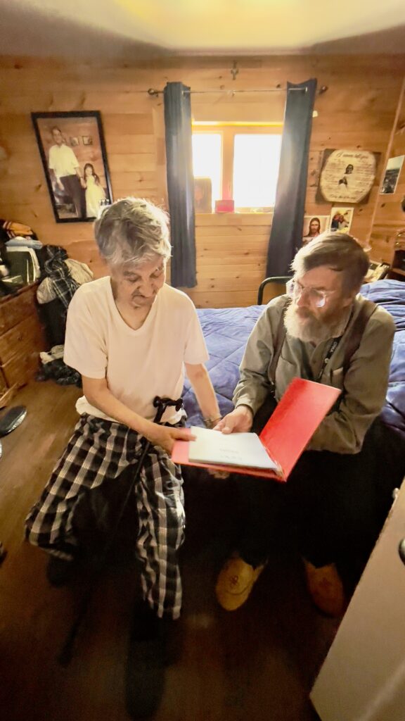 Two elderly men are seated together in a cozy, wood-paneled room. One man, holding a cane, is wearing a white t-shirt and plaid pants, while the other, wearing glasses and a collared shirt, shows him a red folder with papers inside. The room is warmly lit, and family photos and religious images adorn the walls, creating a comforting atmosphere.