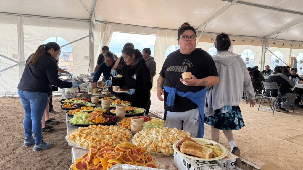 A large breakfast buffet spread inside a tent. Several trays of fruit, cheese, and toast are laid out on a table, with people lining up to serve themselves. A woman in a black "Buffalo" T-shirt holds a cup of food and stands near the buffet table, while others serve themselves in the background. The setting is lively, with multiple people moving around, and tables are filled with attendees enjoying breakfast.