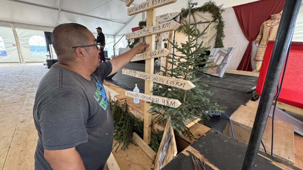 A man, identified as John Chescappio, stands beside a wooden signpost placed on the stage in a large tent. The signpost has directional signs showing the distances from the gathering site to various nearby communities, including Waswanipi (856 km), Masteuiatsh (803 km), and Kawawachikamach (9 km). John is pointing towards one of the signs. The stage is decorated with evergreen branches and traditional items, including a mannequin wearing Indigenous attire. The scene evokes a sense of connection among the communities represented.