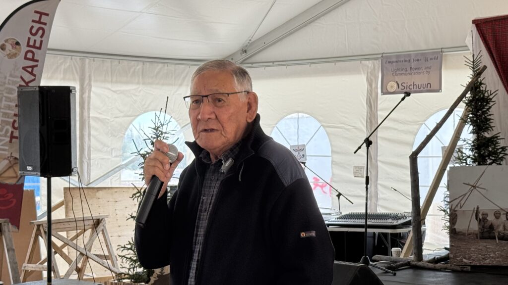 Michapow (Paul Arthur McKenzie) stands holding a microphone, leading the group in prayer before a meal at the Elders Gathering. He is dressed in a dark jacket, with glasses and short silver hair. Behind him, a large tent with curved windows can be seen, decorated with evergreen trees and banners. A microphone stand is nearby, and a banner in the background reads, "Empowering Your World - Lighting, Power, and Communications by Sichuun."