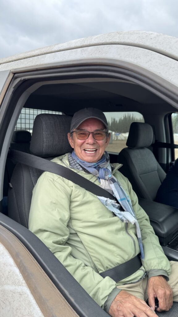 Raoul Vollant, wearing a light green jacket and gray scarf, is sitting in the front seat of a vehicle, smiling broadly through the open window. He appears joyful and ready for his next task at the Elders Gathering.