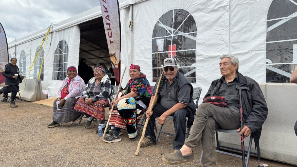 A group of five elders, four women and one man, are seated outside a large white tent at the gathering. They are all dressed in colorful traditional clothing, with the two women on the left wearing bandanas and the man on the far right holding a carved walking stick. They are sitting comfortably and smiling, enjoying their time together.