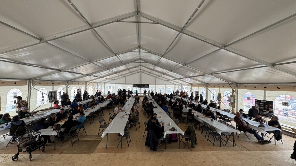 A wide-angle view of the interior of a large event tent, with long rows of tables filled with people. The tables are covered with white tablecloths, and attendees are seated or standing, socializing and eating. The bright, open space has windows along the sides, letting in natural light. The atmosphere is busy and communal, with many elders and families gathered for a meal at the Elders Gathering.