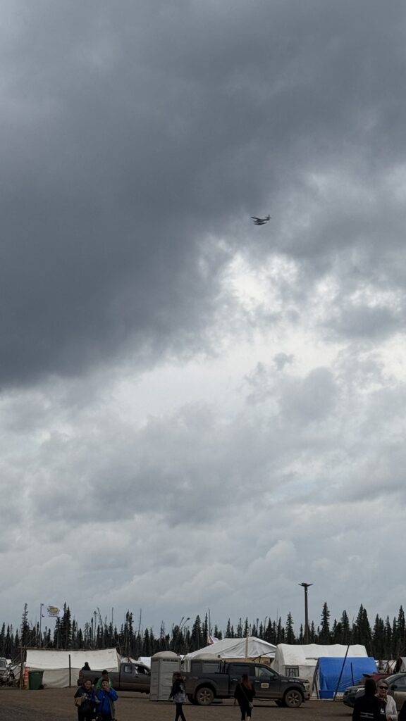 A floatplane flies high above the Elders Gathering camp, with thick clouds in the background. Below, a few people walk around the tents and vehicles parked in the camp area. The tall trees of the northern forest line the horizon, adding a rugged and remote feel to the scene.