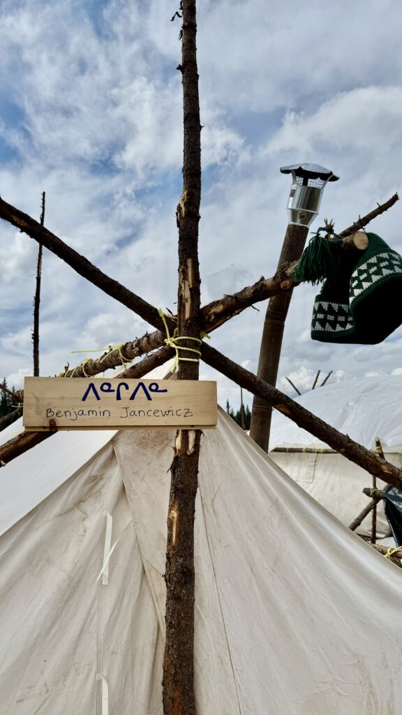 A handmade wooden sign hangs at the entrance of Benjamin Jancewicz's tent. The sign is tied with yellow rope to a simple structure made from branches and features Benjamin’s name written in both Naskapi syllabics and English. A knitted green hat hangs nearby, and a chimney pipe from the tent’s woodstove is visible in the background. The sky is partially cloudy, casting soft light over the camp. The sign is a meaningful, personal touch in an otherwise rustic setup.