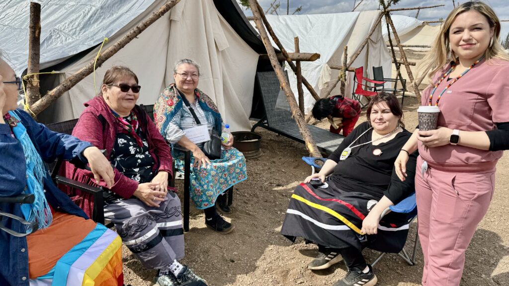 Image of women sitting by tents: A group of women, some wearing traditional skirts and shawls, sit together near a tent at the camp, engaging in conversation. They are gathered around a couple of camping chairs, and the sun is shining, casting a warm light on their faces. One of the women is standing, holding a cup of coffee, while the others are seated.