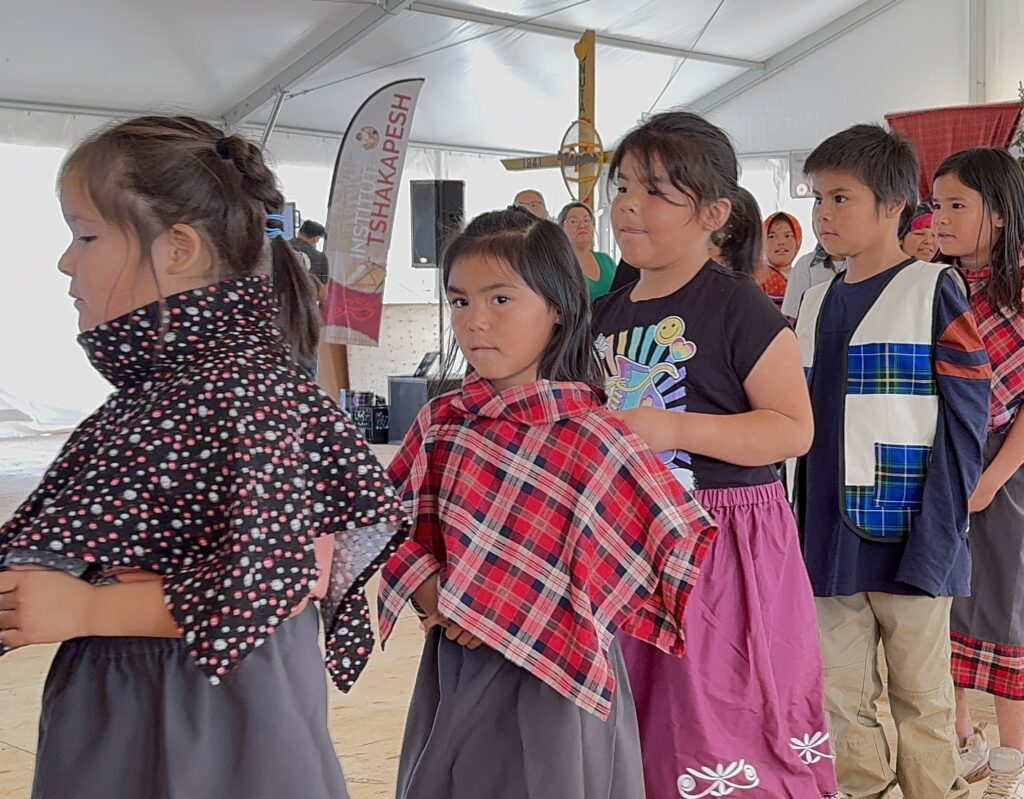 A group of young students from Jimmy Sandy Memorial School's Grade 2 class, dressed in a mix of traditional Naskapi plaid clothing and modern attire, line up for a dance. Their faces show focus and anticipation as they perform at the Opening Ceremony of the event. Some wear plaid capes or vests, with a few looking directly at the camera.