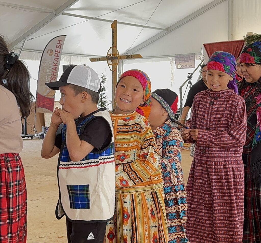 More children from the Jimmy Sandy Memorial School Grade 2 class are seen in traditional clothing. One of the boys wears a white vest with blue plaid panels and a black shirt, while others don bright colors and headscarves. A young girl in a vibrant orange and yellow dress smiles joyfully as the group continues their performance.