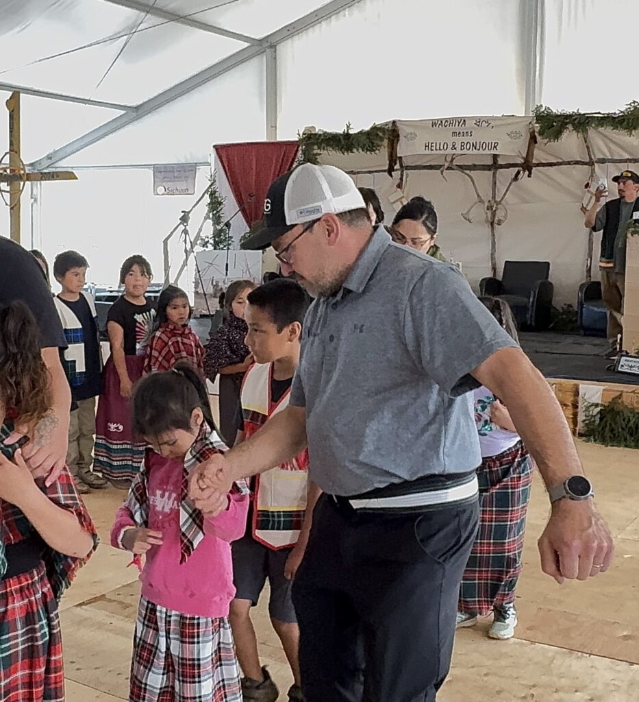 Noah Swappie, wearing a gray polo shirt and a baseball cap, helps guide the students in their traditional dance. He holds the hand of a young girl in a pink hoodie, leading her through the steps while other students in plaid and traditional outfits look on or continue dancing in the background.