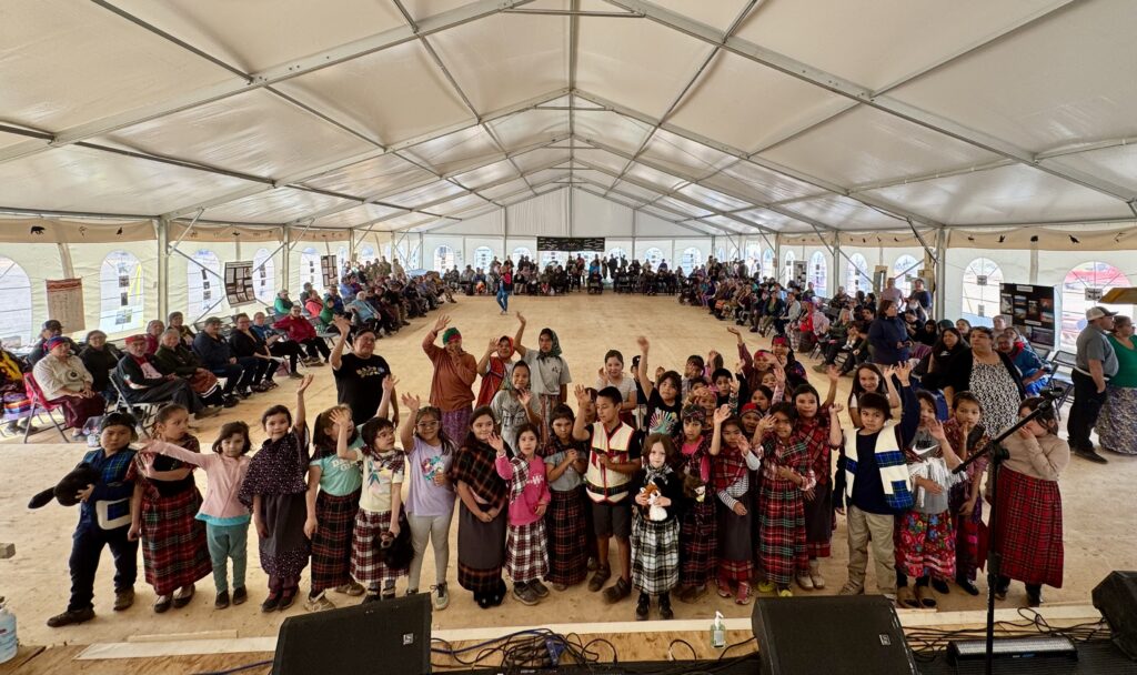 A large group of children stands on stage, facing the camera with many smiling and waving enthusiastically. The kids, some dressed in traditional plaid clothing, are gathered in a semicircle at the front of the stage. Behind them, a large audience sits along the edges of the tent, watching the performance. The tent is spacious, with a high ceiling and large windows letting in natural light. The scene captures the joy and energy of the children's performance during the event.