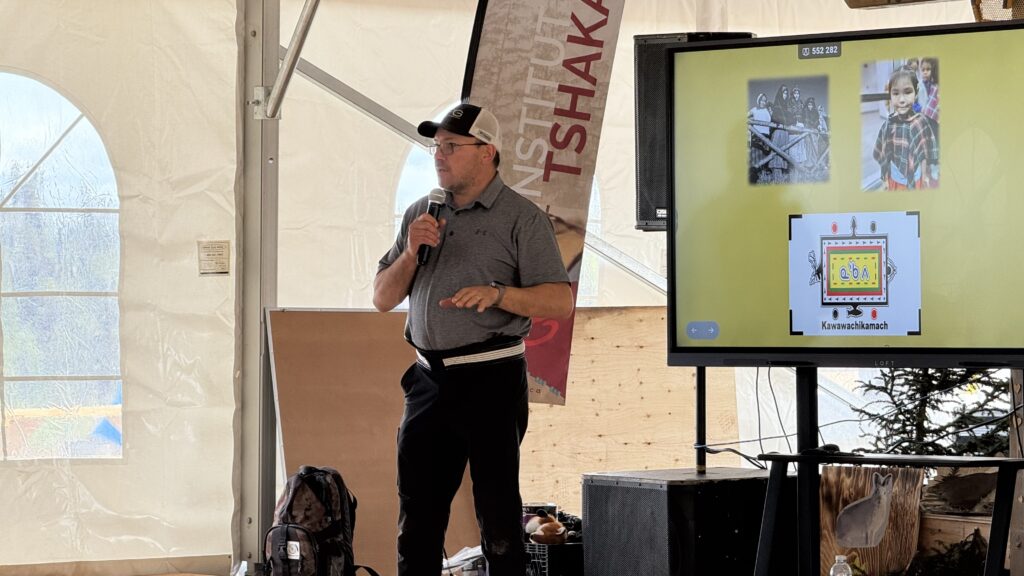 Noah Swappie stands in front of an audience, holding a microphone and presenting a slideshow on Naskapi history. Behind him is a large monitor showing a slide with images of Naskapi people and the Kawawachikamach logo. A vertical banner for the Institut Tshakapesh is visible in the background. He is wearing a gray shirt, black pants, and a hat, with a sash tied around his waist, symbolizing his cultural pride. A backpack sits on the floor near him.