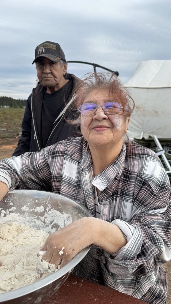 An elder woman smiles as she kneads dough in a large mixing bowl, her hands and face dusted with flour. She wears a checkered flannel shirt and glasses, while a man, likely another elder, stands behind her wearing a black shirt and a baseball cap. They are outdoors near a tent, as preparations for meals continue at the gathering. The expression on her face reflects her focus and warmth despite the busy kitchen setting.