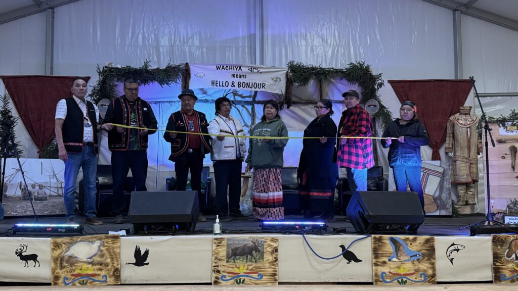 A group of Chiefs and dignitaries from various nations stand together on stage, holding a yellow ribbon, as part of the official opening of the event. Chief Theresa Chemaganish is present alongside others, under a banner that reads "WACHIYA means HELLO & BONJOUR." The stage is decorated with symbols of nature, including a backdrop of spruce branches and traditional Naskapi art.