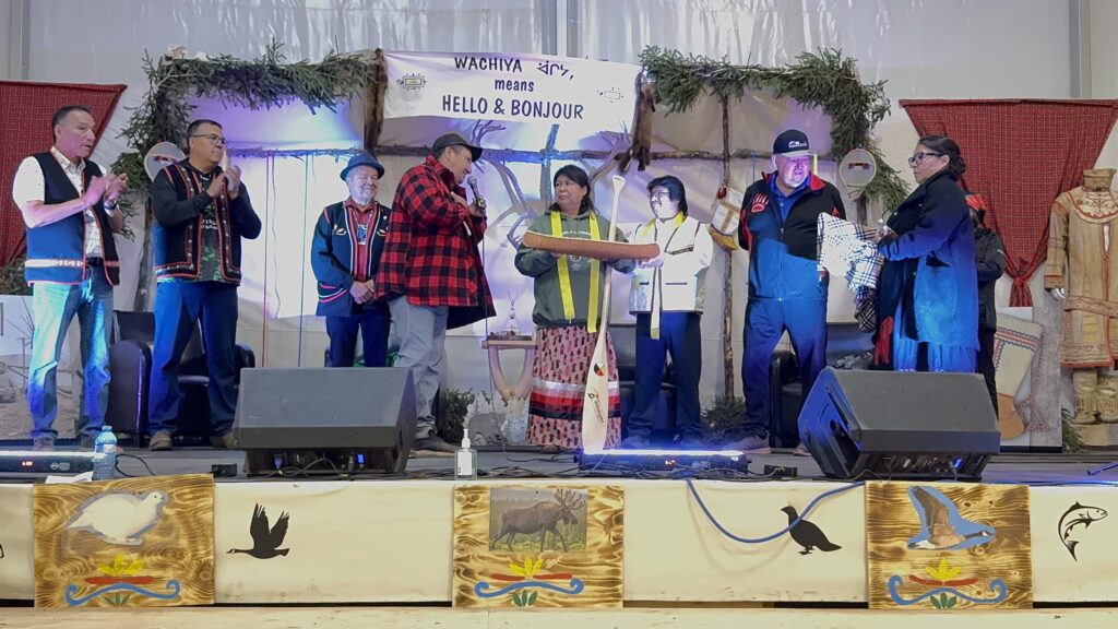 Chiefs and dignitaries from multiple nations gather on stage, presenting gifts to Chief Theresa Chemaganish and event organizers. Chief Theresa holds a ceremonial paddle, while others offer traditional gifts such as blankets. The event takes place under the same decorated stage backdrop, and the gesture signifies the importance of unity and mutual respect among the nations.