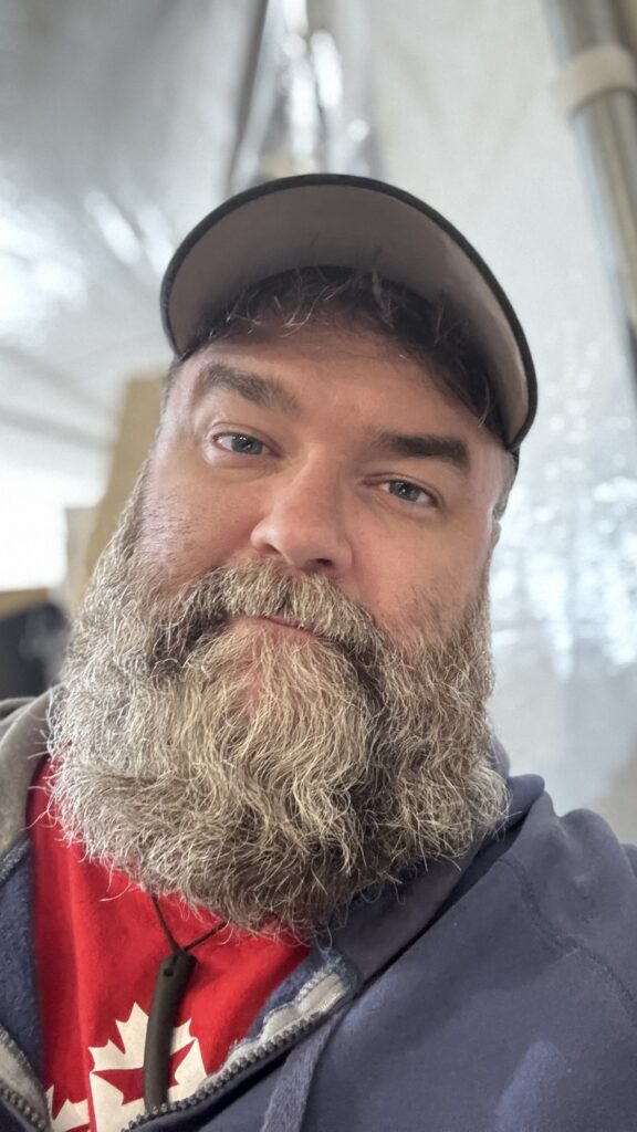 A close-up selfie of Benjamin Jancewicz, showing a clear view of their bearded face, wearing a cap and a red shirt. The expression on their face looks slightly weary but calm, with the background blurred behind them, likely taken in a tent or indoor setting.