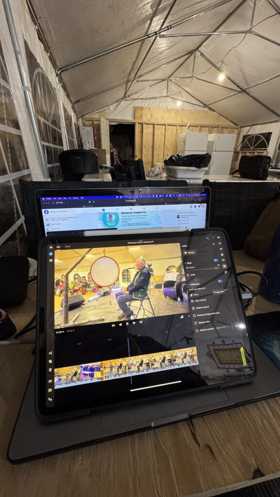 A makeshift video editing station set up inside a large tent. An iPad is open on top of a laptop, showing a video editing interface with a scene of Michapow drumming. Various cables, chargers, and devices are visible, indicating a busy workspace. The background shows a kitchen area with refrigerators and dim lighting.