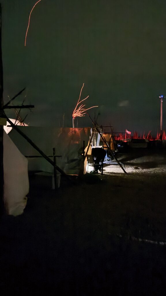 A night shot of a campsite, with tents made from canvas and wooden structures visible under a dark sky. Glowing embers from a fire shoot upward, creating streaks of orange light. A small spotlight in the distance adds a cool blue hue to the scene.