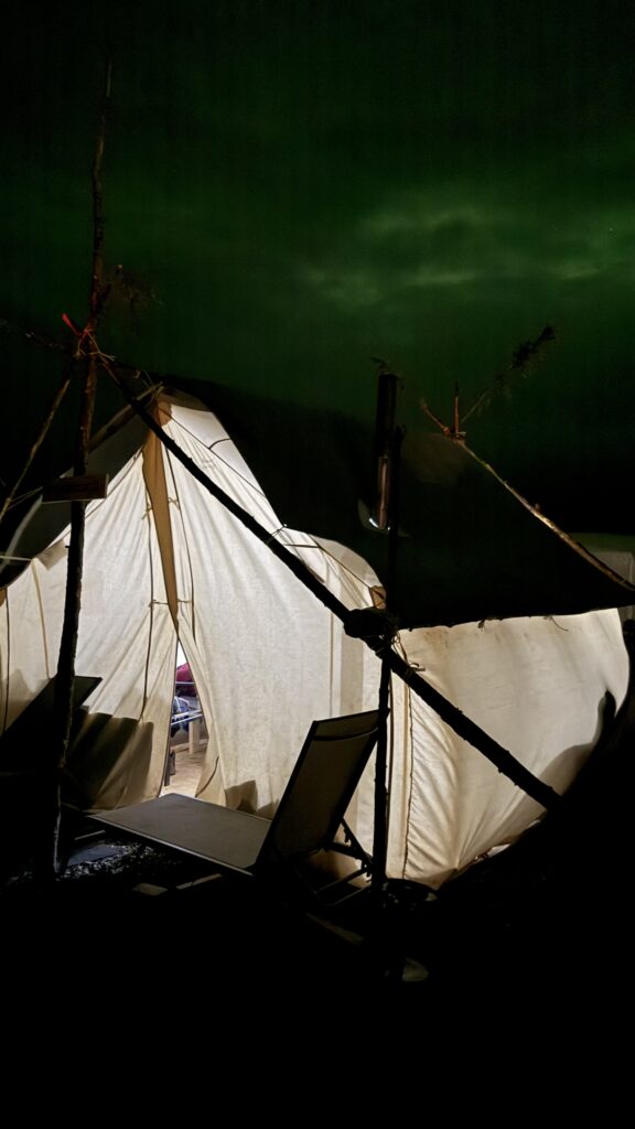 Another night view of the campsite, this time focusing on a well-lit tent entrance, illuminated from within. A chair leans against the frame of the tent, while the greenish hue of the Northern Lights can be faintly seen in the sky above.