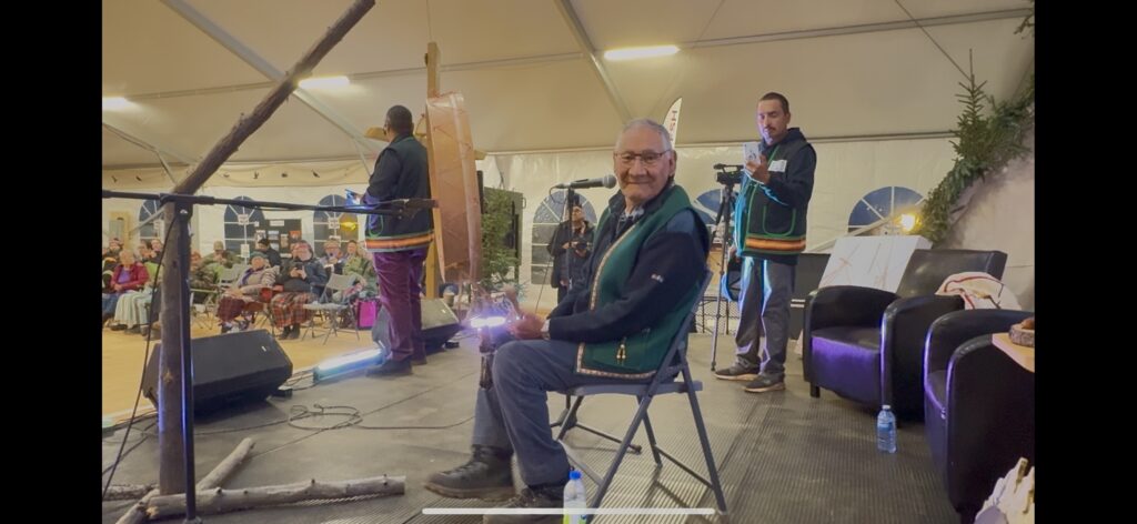 Michapow (Paul Arthur McKenzie) is seated on stage, holding drumsticks and smiling at the camera, preparing to start the Opening Ceremonies with traditional drumming. Behind him, other men in traditional vests are seen preparing and filming the event. The audience is seated in the background, wrapped in warm clothing and blankets, watching attentively. The setting is within a large, warmly lit tent adorned with Naskapi art and decorations.