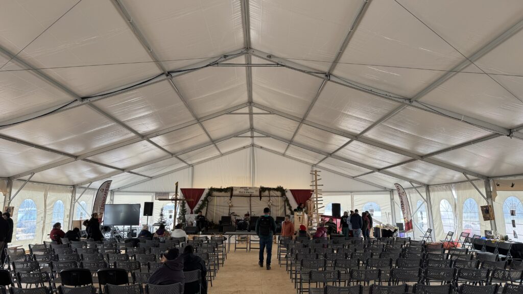 A view of the large event tent set up for the gathering. Rows of empty chairs are positioned in the foreground, facing a stage decorated with greenery, drums, and a sign that reads "Wachiya means Hello & Bonjour." A few people are seen standing near the stage and scattered throughout the tent. The event banners and various displays line the tent's sides.