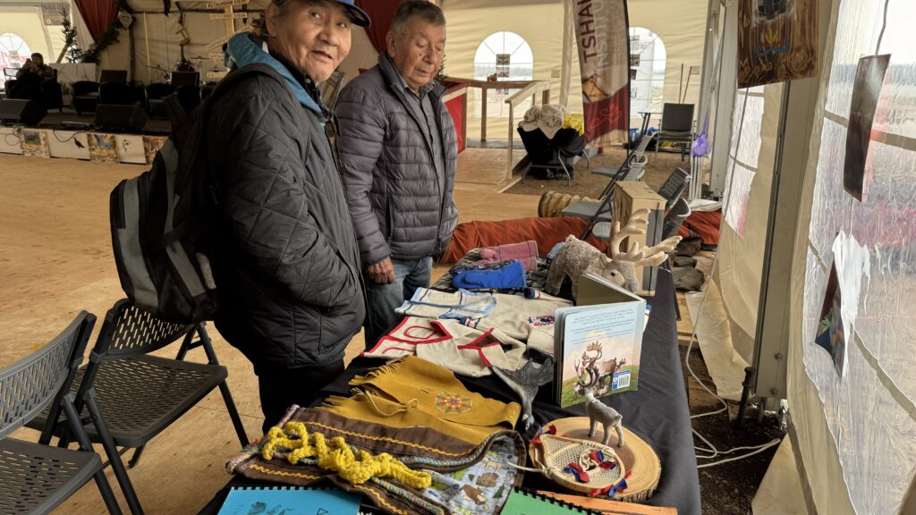 Two elders stand next to a display table filled with Naskapi artifacts and crafts at the Naskapi Development Corporation exhibit. The table showcases traditional items, including a beaded pouch, a small reindeer figurine, and a colorful fabric piece. The elders appear to be appreciating the cultural items, reflecting the rich history of the Naskapi people. In the background, a stage and more seating are visible under a large event tent.