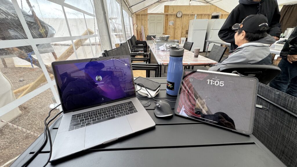 A small workstation set up in the kitchen tent includes a laptop, mouse, iPad, and water bottle on a black table. The laptop displays a login screen, while the iPad is locked, showing the time as 11:05. In the background, a long table with kitchen equipment, including a large bowl and mugs, is visible. A person wearing a "Native Pride" cap sits nearby. The reflective surfaces capture the photographer's reflection, adding a personal touch to the thoughtful breakfast setup arranged for them.