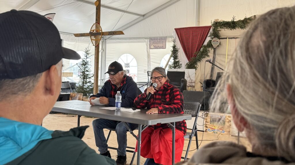 Caroline St-Onge speaks into a microphone while leading a session on grief. She is sitting at a table alongside another elder, both dressed in warm clothing. The backdrop includes a large cross, banners, and festive greenery decorating the tent.