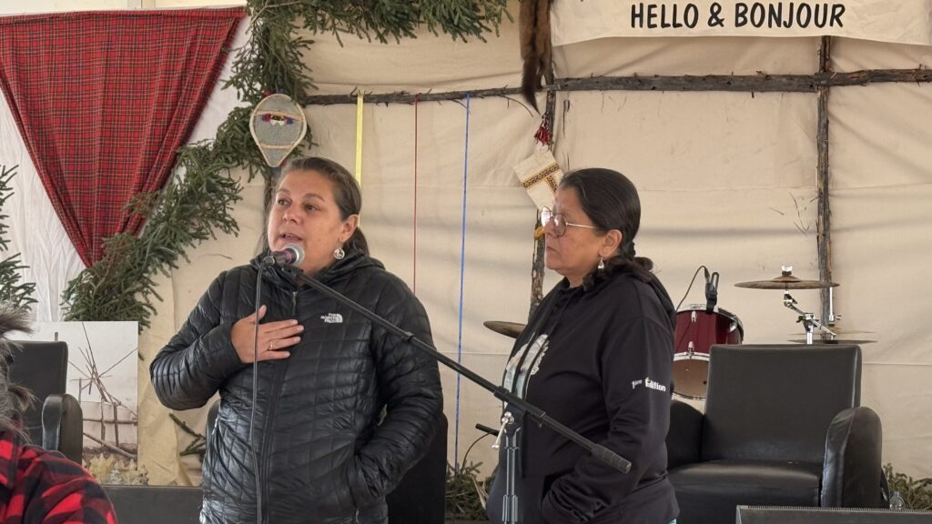  A woman in a black jacket, standing at a microphone, speaks with her hand on her chest. Another woman stands beside her, listening attentively. Both women are part of a session held under a large tent with traditional decor, including greenery and woven crafts.