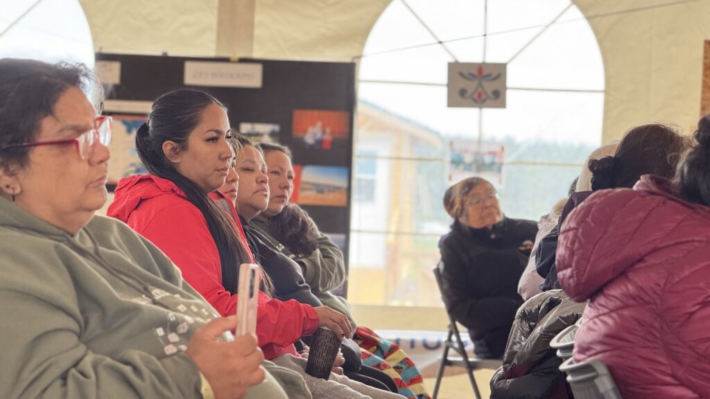 A close-up shot of several women sitting in rows during the grief session. They listen intently, with somber and reflective expressions. The tent’s arched windows and display boards can be seen in the background.