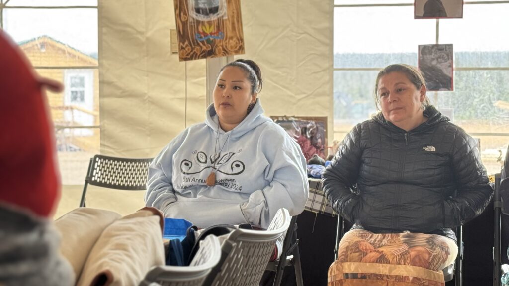 Two women sitting in the audience during the grief session, both dressed warmly. One woman wears a light blue hoodie with a Naskapi event logo, while the other is wearing a black jacket. They sit quietly, processing the emotional moment.
