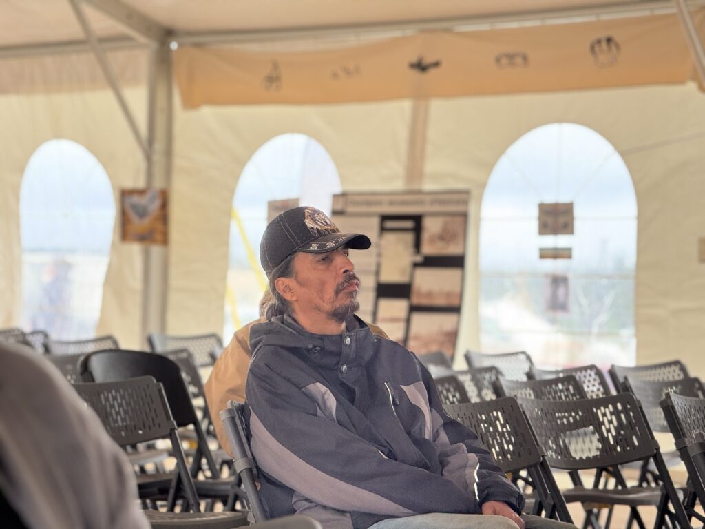 A man, seated alone, listens thoughtfully during the grief session. He wears a black cap and a gray jacket. Behind him, empty rows of chairs and a decorated wall with various posters and displays are visible.