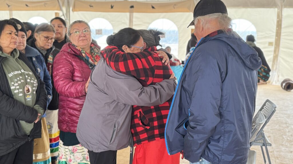 Two women embrace each other tightly, offering comfort during the grief session. Both are wearing patterned jackets. Other members of the community, also visibly emotional, stand nearby in the background.