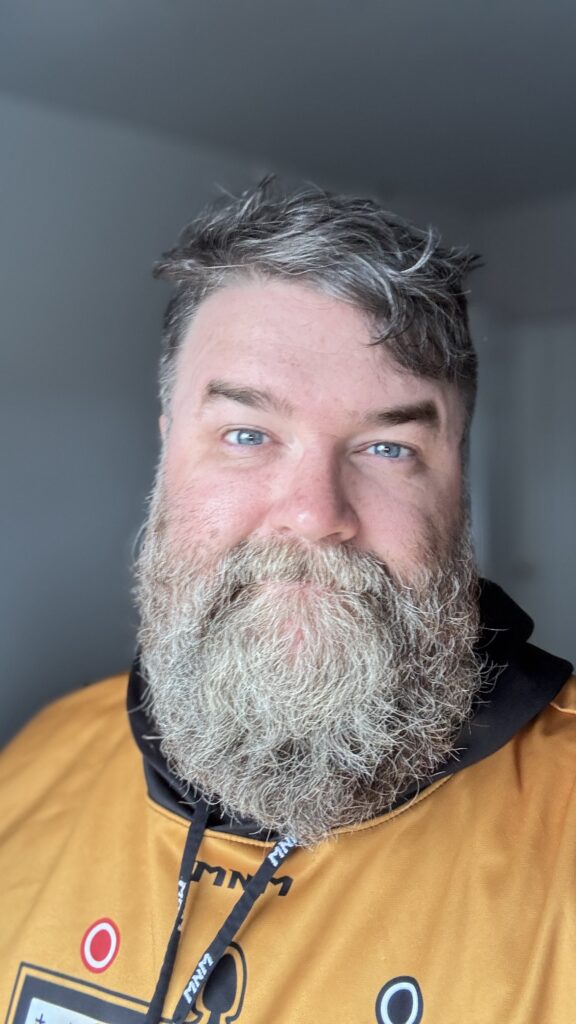 A close-up selfie of Benjamin Jancewicz wearing a cap and a gold-colored hoodie. His beard is thick and graying, and his expression is neutral, with a slight smile.