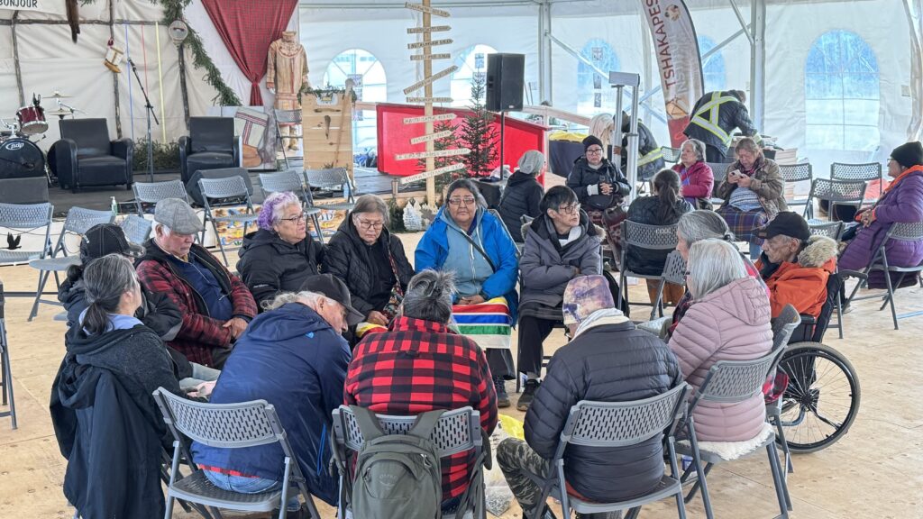 A circle of elders sits together under a large tent. The group is deep in conversation, some wearing colorful blankets and hats. Behind them, the stage is decorated with traditional symbols, and several other people are seated around the tent, engaging in their own conversations.