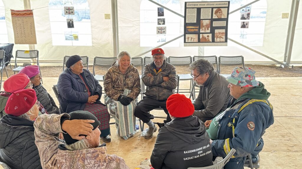 A second group of elders sits in a sharing circle under the same large tent. This group is also engaged in conversation, with one elder draped in a colorful Hudson’s Bay blanket. Posters displaying historical information are visible on the tent wall behind them.