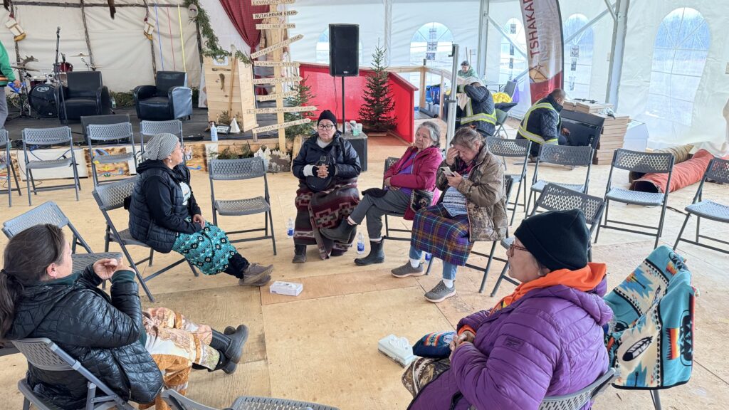 A smaller group of women sits in a circle, having a discussion under the large tent. One woman wears a blanket in traditional colors, and others are bundled up in warm clothing, indicating a chilly environment.