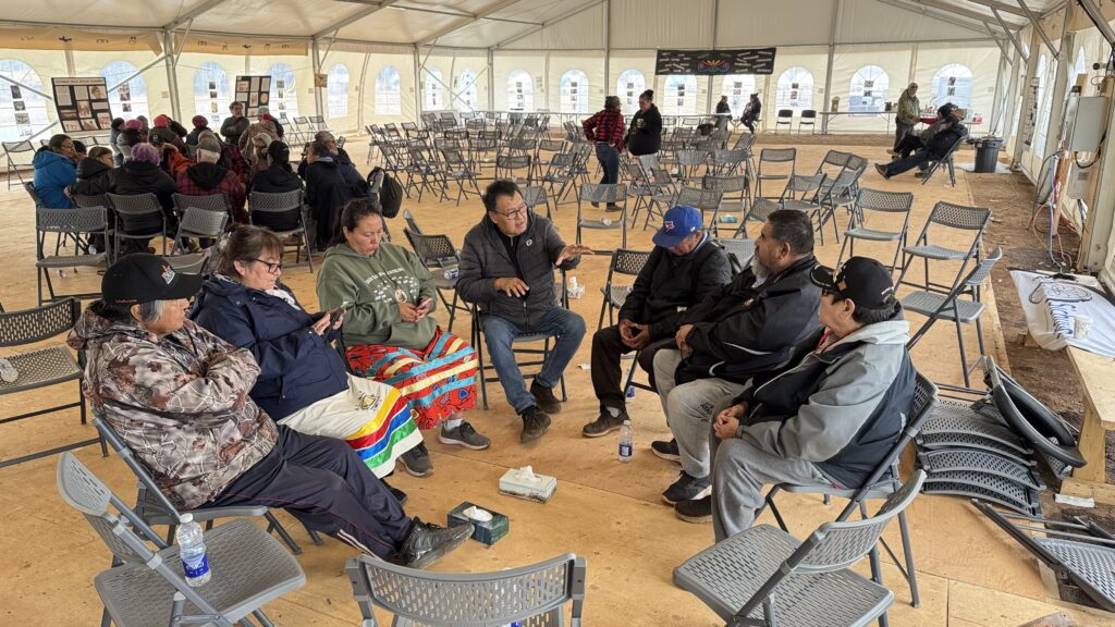 A group of men and women sits in a sharing circle, talking intently under the large tent. The man leading the conversation gestures with his hands, while the others listen. The gathering is peaceful, with some people observing from a distance.