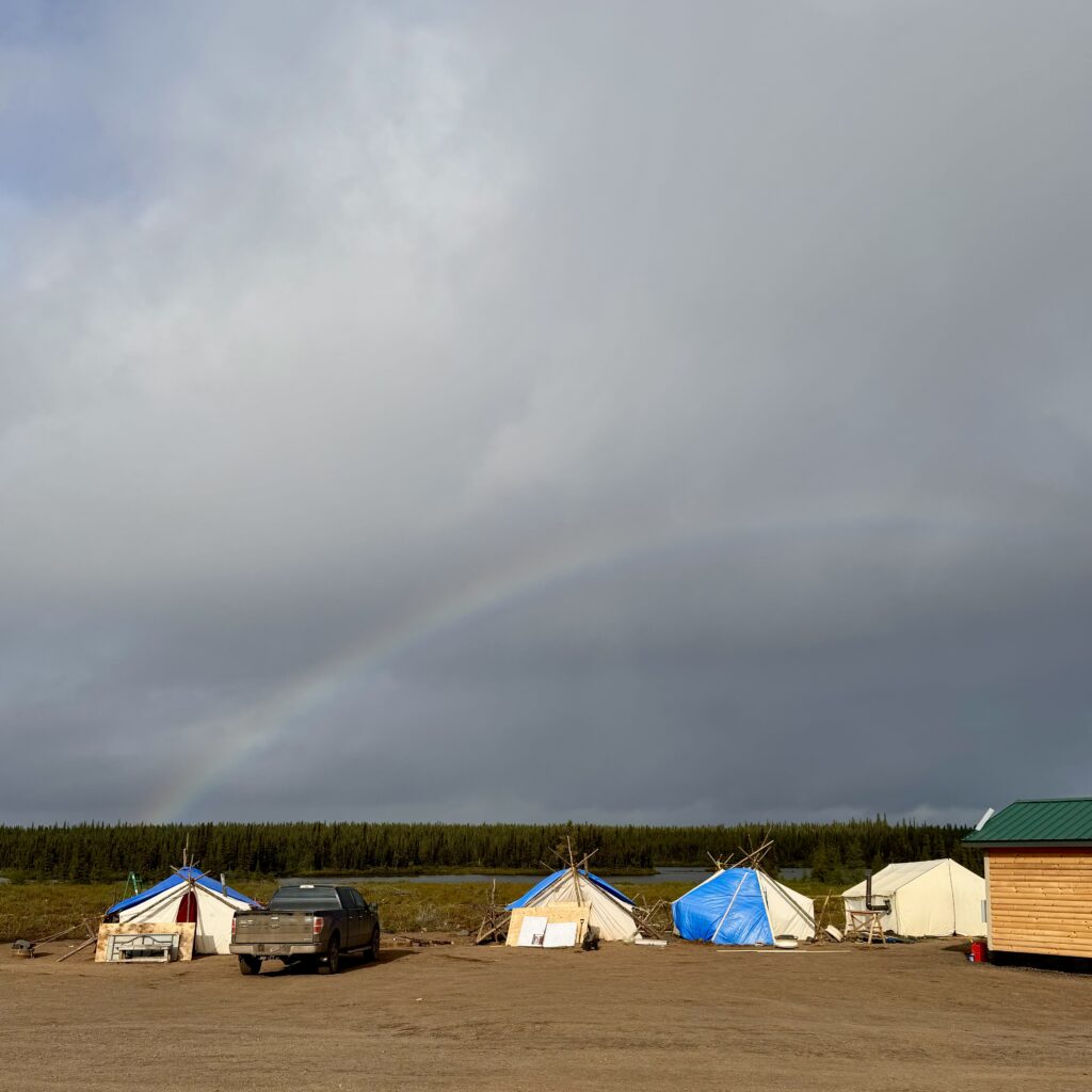 A scene featuring a wide rainbow arcing over several tents in a camp. The tents are constructed with wooden frames and some are covered with tarps. A dark truck is parked nearby, and the sky is a mix of rain clouds and clearing blue patches.