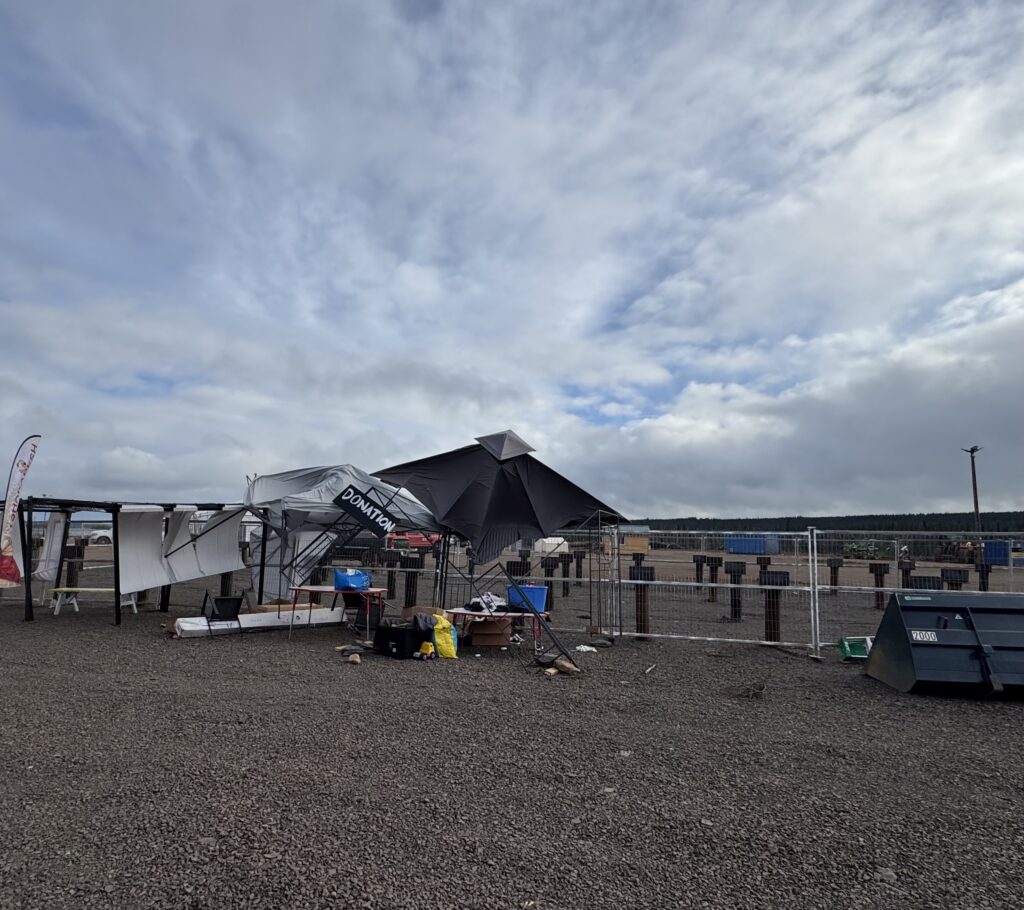 A collapsed donation center tent in a gravel lot, with poles and fabric scattered by the wind. Behind it, there are other tents and a large field extending into the distance under a mostly cloudy sky.