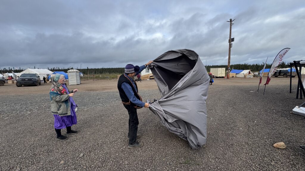 A man wearing a blue knit hat and blue vest struggles to fold a large gray tarp in the wind, with an older woman in a purple dress and patterned shawl standing nearby. They are on a gravel lot with tents and utility poles visible in the background, and both seem to be working to salvage materials after the storm.