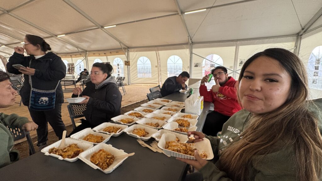 A group of people sitting around a table in a large tent enjoying a meal together. Several take-out containers filled with lasagna are spread across the table. One person is giving a thumbs-up while holding a soda can, and a young woman in the foreground is eating. The atmosphere feels communal, with everyone taking a well-deserved break from their busy day.