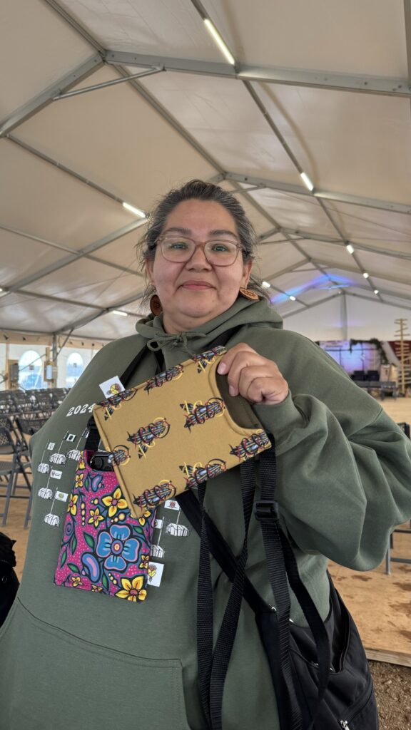 Cheyenne, standing inside the large tent, smiling and holding a beautiful handmade phone bag. She is wearing a green hoodie with a colorful design that matches the bag’s feathers. Her sense of pride and joy is evident as she presents the gift, which she generously distributed to the workers. The tent in the background is set up for the event, with chairs and tables arranged.