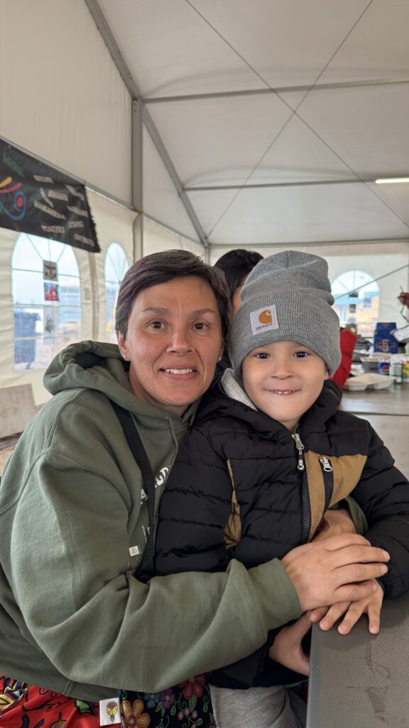 A woman wearing a green hoodie with colorful floral designs smiles warmly as she embraces a young boy sitting on her lap. The boy is bundled up in a black and tan jacket and wears a gray Carhartt beanie, smiling slightly. They are seated at a table inside a large tent with bright, natural light streaming in from the windows. The closeness and joy shared between them reflect the familial warmth and the importance of community at the elders' gathering.