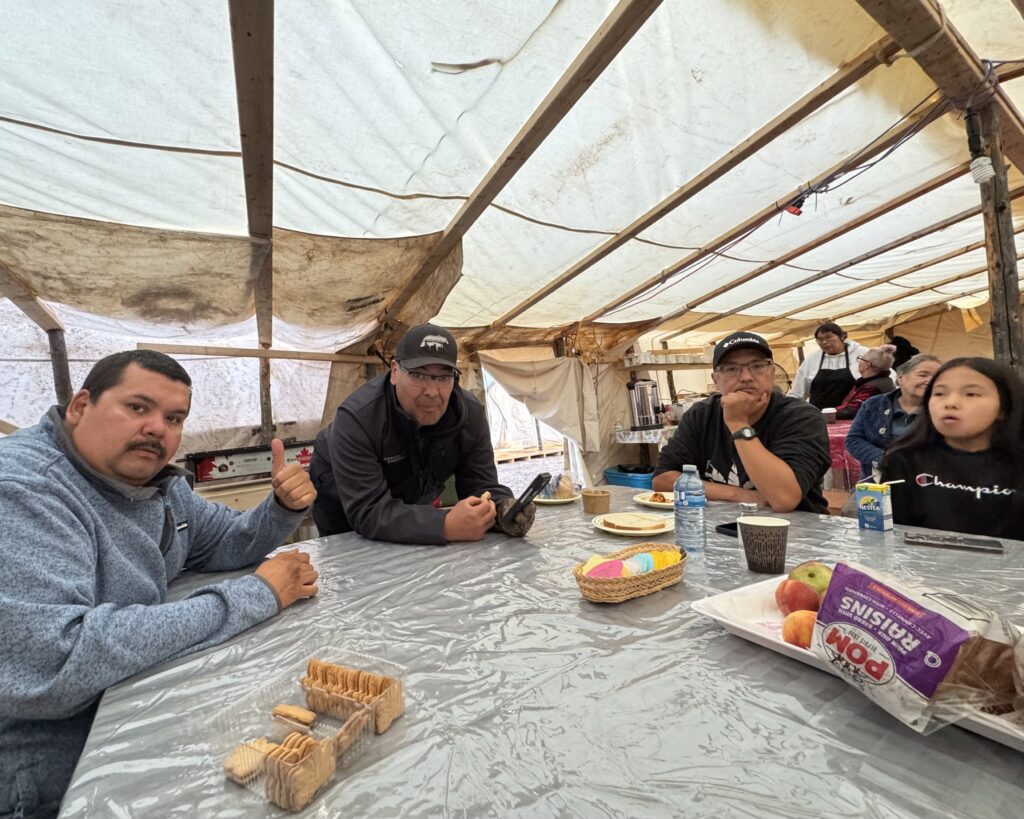 Inside a cozy Innu kitchen tent, four people sit around a table covered with a plastic tablecloth, enjoying a break. A man in a blue sweater gives a thumbs-up while another in a black jacket with glasses looks thoughtfully. To the right, a man in a black hoodie and cap sits with a hand on his chin, and a young girl with long dark hair in a black Champion sweatshirt looks off to the side. On the table are cookies, apples, a box of raisins, and other snacks. The tent's rustic wooden beams and tarps provide warmth and shelter, creating an atmosphere of shared stories and comfort.