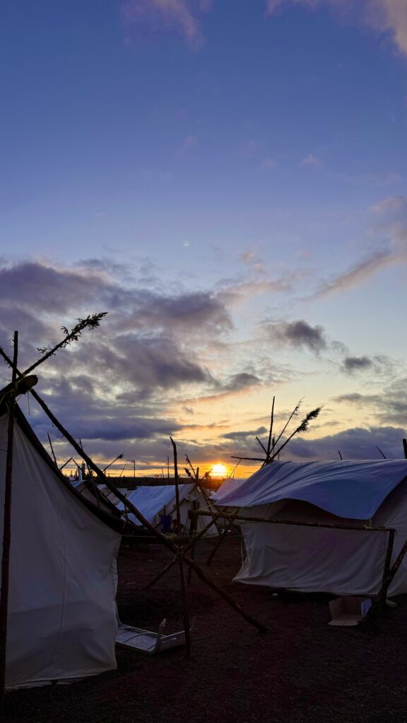 A stunning sunset over a camp with several white tents. The tents are supported by wooden poles, with branches extending from the structures. The sky is a mix of deep blue and purple hues, with clouds glowing orange from the setting sun. The scene feels serene and peaceful as the day transitions into night.