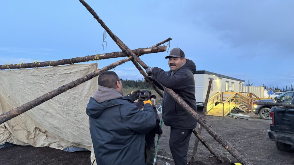 Two men are working together to secure wooden poles for a tent. One man, smiling, holds the crossbeam while the other secures the structure with yellow ropes. The scene takes place outdoors near a construction area, with portable buildings and vehicles in the background under the evening sky.