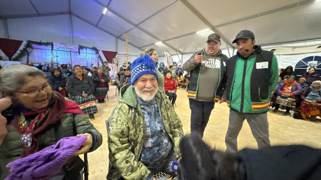 A man in a blue knitted hat and camo jacket smiles as he sits in the front row of a gathering tent. A woman next to him hands him a purple knitted item, while others in the background watch attentively. Two men, one holding a microphone, stand nearby, preparing for an announcement.